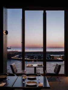 A modern restaurant interior with a sea view, featuring wooden tables and chairs, plates, and glasses set on a table.