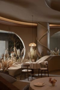 A modern restaurant interior with neutral textiles, featuring round tables, chairs, and decorative plants.