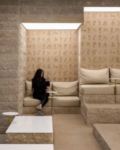 A minimalist cafe interior with beige concrete walls and floors, featuring a woman sitting on a cushioned bench with a smartp