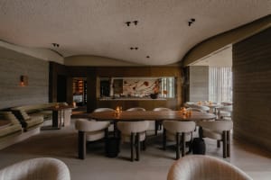 Modern restaurant interior with wooden tables, fabric upholstered chairs, and a bar area with a metal counter.