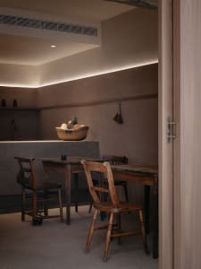 A minimalist residential kitchen with wood and concrete materials, featuring a basket of fruits and a vintage table and chair