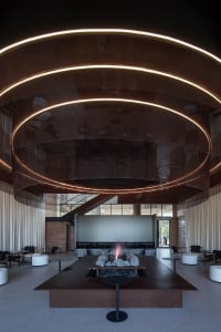 An industrial-style lobby with brick walls, metal railings, and a glass ceiling, featuring a central fire pit.