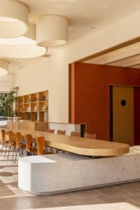 A modern cafe interior featuring a long wooden bar counter with terrazzo base, orange chairs, and a red accent wall.