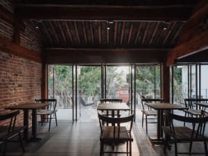Industrial-style cafe interior featuring wooden tables and chairs, brick walls, and large windows overlooking greenery.