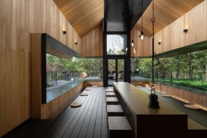 A minimalist restaurant interior with wooden tables and benches, surrounded by large windows overlooking greenery.