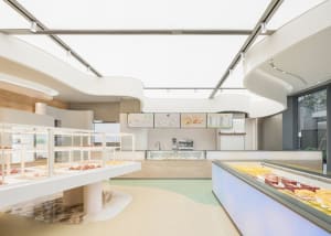 A modern bakery with white concrete interior features shelves of pastries and a counter displaying cakes.