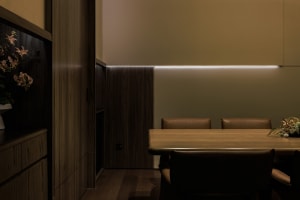 A minimalist dining area with wooden table and leather chairs, illuminated by hidden lighting under the wall.
