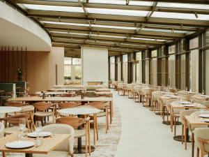 A modern restaurant interior with wooden tables, chairs, and glass panels, featuring a spacious and airy atmosphere.