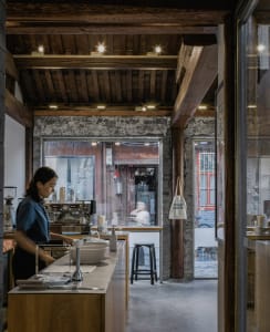 A worker in a blue apron preparing coffee behind a wooden counter in an industrial-style coffee shop with exposed brick and c