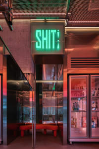 An illuminated industrial neon sign with the word 'SHIT!' in a metallic-framed facade.