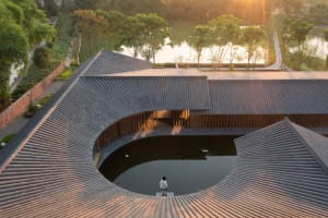 A person standing on a curved clay-tiled roof overlooking a pond and greenery.