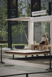 A minimalist urban cafe with wooden platforms, metal seating, glass walls, and fabric curtains.