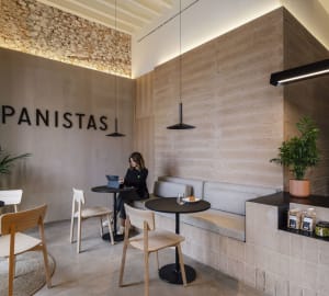 A minimalist cafe with concrete and wood materials, featuring seating, tables, and a wall inscription that says "Panistas."