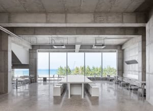 Interior view of a modern restaurant featuring concrete walls, a large window overlooking the beach, and minimalist furniture