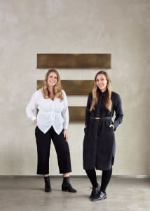 Two women wearing white and black outfits standing against a white wall with gold rectangular panels.