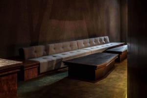 A modern, cushioned lounge bench in a dark, textured room with wooden tables.