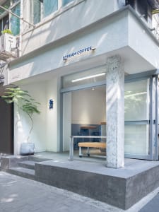 Concrete exterior of a coffee shop with glass doors and a simple seating area inside.