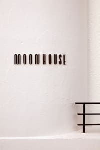 A minimalist storefront with a textured white wall, black railing, and large text reading "MOONHOUSE"