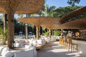 An outdoor lounge area with bamboo ceiling structures and white seating areas surrounded by natural greenery under a clear bl