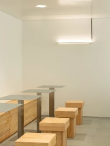 A minimalist interior featuring wooden stools and metal tables on a grey floor.