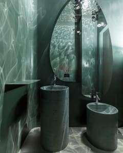 A modern industrial bathroom featuring concrete sinks, a large oval mirror, and glass shelving.