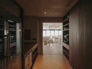 A modern industrial kitchen with wooden cabinets and a concrete floor, featuring a view into a seating area with natural ligh