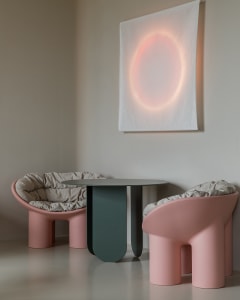 A modern lounge area featuring two pink, curved-plastic chairs with beige cushions and a small, round glass table in a minima
