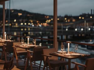 An elegant restaurant setting with dinner tables overlooking a illuminated harbor at dusk.