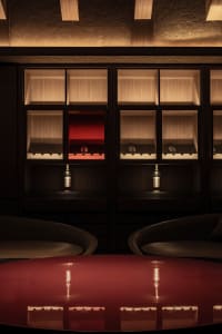 A modern restaurant bar with wooden walls, glass shelves, and a red table with bottles of wine.