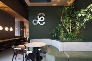A modern industrial coffee shop interior with green wall, curved seating, and potted plants.