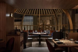 A modern restaurant interior with a combination of concrete and wood materials, featuring round tables and comfortable seatin