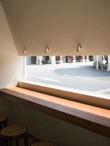 A minimalist coffee shop interior features a wooden window bench with a view of the street.