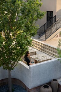 Person sitting at a wooden table in an outdoor lounge area with a concrete wall and tree.