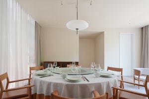 A modern dining room with a round glass-top table, wooden chairs, and transparent windows.