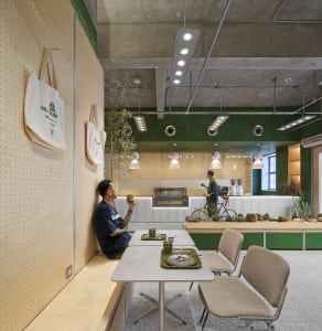 A person sitting at a table in an industrial-style coffee shop with greenery indoors, concrete ceiling, and metal and wood fu