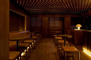 A minimalist restaurant bar with wooden furniture and walls, featuring small round tables and stools.