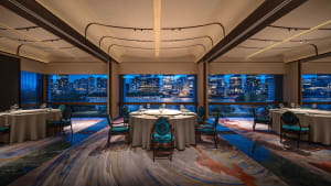 A modern restaurant with blue upholstered chairs, glass windows overlooking a cityscape, and a carpeted floor.