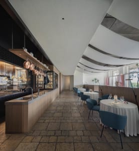 Interior of a modern restaurant with concrete flooring, white tables, blue chairs, and a sleek kitchen counter.