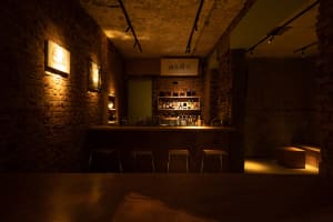 An industrial-style bar with brick walls, featuring a wooden counter and stools, illuminated by warm lighting.