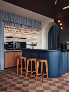 A modern bar with wooden stools, a blue curtain backdrop, and multiple glassware hanging above the counter.