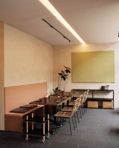 A minimalist restaurant interior featuring wooden tables with woven chairs and concrete accents.