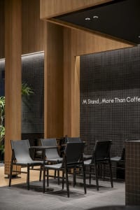 Indoor coffee shop setting with industrial design elements, featuring wooden columns, black metal chairs, and a tiled wall.