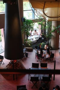 People sitting at tables in a coffee shop with a large column and fire pit, surrounded by plants.