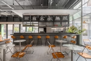 A modern cafe with a metal bar counter featuring sleek glass tables and vibrant yellow seats.