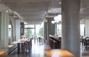A spacious, modern-industrial restaurant interior featuring concrete, glass, and wooden materials.