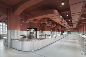An industrial-style cafeteria with a long metal counter, white tile backsplash, and exposed ventilation systems.