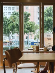 A cafe interior with modern wooden tables and chairs, featuring a window view of trees and buildings.
