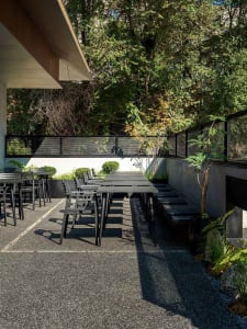 Modern minimalist outdoor dining area with concrete flooring, gray table, and black chairs surrounded by lush greenery and a 
