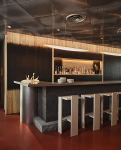 A modern bar with wooden walls and stools, featuring a counter with bottles and plates.
