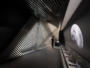 A person standing in front of a metallic wall with slanted metal strips, featuring vintage cameras and a large moon image.